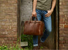 Man leans against a brick wall holding a brown leather briefcase.